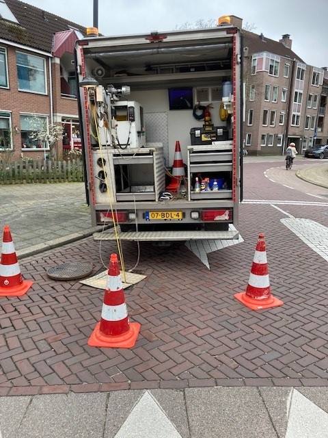 Vrachtwagen in een straat met een kabel die de rioolput in gaat om opnames te maken. De plek is afgezet met pionnen.