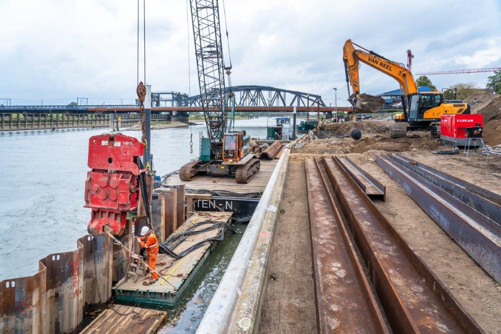 De lange damwanden worden een voor een diep de bodem van de rivier ingeduwd.